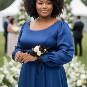 May include: A person in a long-sleeved, navy blue dress with a scoop neckline. They are wearing a wrist corsage of white and black flowers. The background shows a wedding ceremony with white floral arrangements.