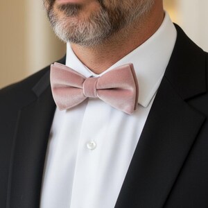 May include: A dusty rose velvet bow tie is the focal point of this image. The bow tie is set against a white dress shirt and a black suit jacket. The image is a close-up, highlighting the texture and color of the bow tie.