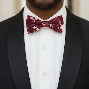 May include: A burgundy bow tie with a floral pattern is the focal point of this image. The bow tie is set against a white dress shirt and a black tuxedo jacket. The floral pattern includes small white and pink flowers.