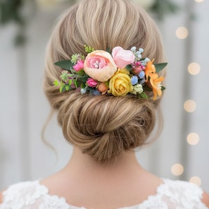 May include: A hair accessory featuring an arrangement of artificial flowers in shades of peach, yellow, pink, and orange, accented with small blue blossoms and green leaves, secured into a blonde updo hairstyle with a white lace garment.