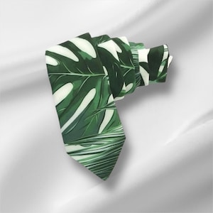 May include: A rolled-up necktie featuring a vibrant green and white tropical leaf pattern. The tie's design showcases large, stylized monstera leaves, creating a bold and eye-catching accessory. The tie is made of a smooth fabric.