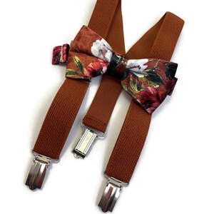 May include: Brown suspenders with a floral bow tie. The bow tie features a pattern of red, white, and green flowers on a brown background. The suspenders have silver-colored metal clips.