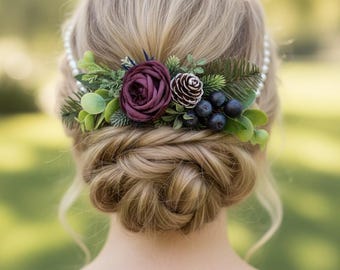 May include: A hair accessory featuring a deep burgundy rose, a small pinecone, dark blue berries, and green foliage. The floral arrangement is attached to a white pearl headband, positioned above a braided blonde hair bun.