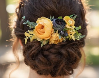 May include: A hair accessory featuring a cluster of artificial yellow peonies, a dark navy blue flower, small dark berries, and green foliage. The floral arrangement is secured into a dark brown braided updo hairstyle with a pearl necklace visible.