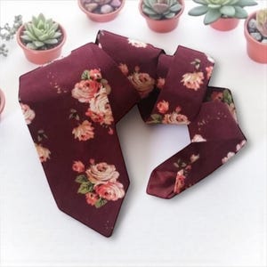 May include: A burgundy necktie with a floral pattern featuring pink and peach roses and green leaves. The tie is made of a smooth fabric and is displayed on a white surface. The tie is a classic accessory.