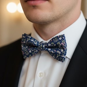 May include: A navy blue floral bow tie with white and pink accents. The bow tie is worn with a white dress shirt and a black suit jacket. The floral pattern adds a touch of elegance to the formal attire.