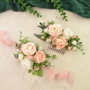 Puede incluir: Dos accesorios florales para el cabello. Uno es un peine con rosas rosas y blancas, vegetación y un peine plateado. El otro es un ramillete de muñeca con flores similares y una cinta rosa. El fondo es una superficie texturizada blanquecina.