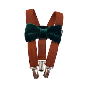 May include: A set of brown suspenders with a dark green velvet bow tie. The suspenders have silver-colored metal clips and a brown leather-like connector. The bow tie is centered on the suspenders.