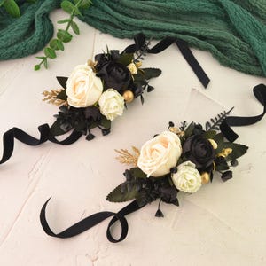 May include: Two floral wrist corsages with black ribbons. Each corsage features cream and black roses, gold accents, and dark green leaves. The ribbons are long and flowing, creating an elegant look.