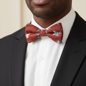 May include: A rust-colored bow tie with a floral pattern in light blue and white is the focal point of this image. The bow tie is set against a white dress shirt and a black suit jacket. The image is a close-up of the upper body.