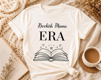 Pode incluir: Camiseta creme com a frase "Bookish Mama ERA" acima de uma ilustração de livro aberto com estrelas e corações. A camisa é amarrada na parte inferior. Uma xícara de café e um livro aberto estão no fundo.