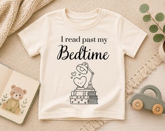 Pode incluir: Camiseta creme de manga curta com o texto "I read past my Bedtime" em escrita preta. Abaixo, um gráfico de uma lâmpada em forma de coração em cima de uma pilha de livros. A camisa é feita de material macio.