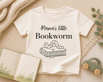 Pode incluir: Camiseta creme com o texto "Mama's little Bookworm" e uma ilustração de desenho animado de um bicho-de-conta. Um livro aberto e um carro de brinquedo de madeira também são visíveis.