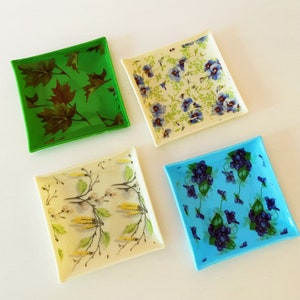 May include: Four square glass dishes with decorative floral and leaf designs. The dishes are in shades of green, cream, and blue. One dish features brown leaves on a green background. Another has blue flowers on a cream background.