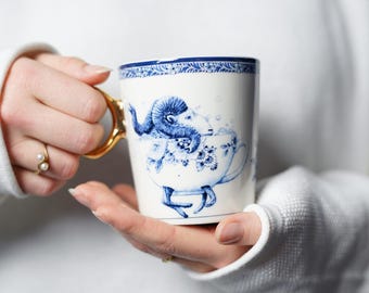 May include: A white ceramic mug with a blue and gold design. The mug features a ram in a bathtub with blue and white floral accents. The mug has a gold handle.