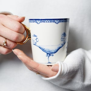 May include: A white ceramic mug with a blue and gold design. The mug features a bird with a long neck and a detailed design. The mug has a gold handle.