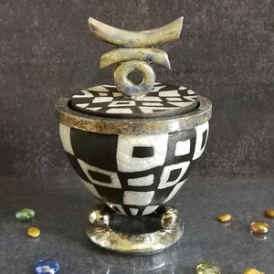 Peut inclure: Un pot en céramique noir et blanc avec un motif géométrique et un couvercle argenté. Le couvercle est surmonté d'un motif abstrait argent et bleu. Le pot est posé sur une surface gris foncé.