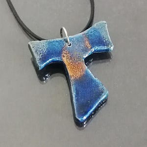 May include: A blue and orange pendant necklace with a T-shaped design. The pendant is suspended from a black cord. The pendant has a glossy finish and a metallic sheen. The necklace is displayed on a dark gray surface.
