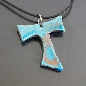 Puede incluir: Un colgante en forma de cruz Tau con un acabado craquelado en tonos turquesa y cobre. El dije en forma de T cuelga de un cordón negro sencillo mediante una argolla de metal plateado, mostrando una superficie texturizada.