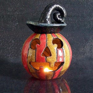 May include: A ceramic jack-o'-lantern candle holder featuring vertical red and yellow stripes, a carved face with cutouts for eyes, nose, and mouth, and a glossy black witch hat topper. A small light glows from within the hollow pumpkin body.