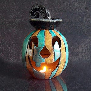 May include: A ceramic jack-o'-lantern candle holder featuring vertical stripes in shades of teal and tan. The pumpkin has a carved face with triangular eyes and a jagged mouth, topped with a black, curled, hat-like stem. A warm light glows from within.
