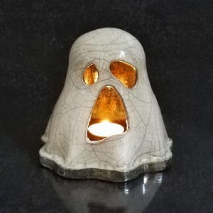 May include: A ceramic ghost-shaped candle holder with a lit candle inside. The ghost is white with gold-colored eye and mouth openings. The candle holder has a crackle glaze finish and sits on a dark surface.
