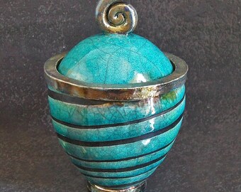 Puede incluir: Un tarro de cerámica turquesa con tapa y asa en espiral. El tarro tiene rayas horizontales negras y ribetes plateados. La tapa es de un tono turquesa más claro y tiene una textura craquelada. Un artículo decorativo.