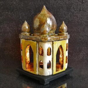 Peut inclure: Une réplique miniature et artisanale du Taj Mahal. Le modèle présente un toit en dôme et de petites flèches, avec des fenêtres cintrées et une palette de couleurs jaune et blanc. Il repose sur une base noire, se reflétant sur la surface brillante en dessous. Le design est détaillé et orné.