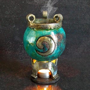 May include: A round ceramic incense burner with a teal and metallic finish, featuring a spiral design on the front and two small handles at the top. The burner sits on a metallic base containing a lit tea light candle emitting smoke.