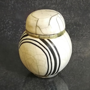 May include: A small, round, lidded ceramic jar with a white crackle glaze. The jar features three bold black stripes and a matching lid. The jar is a decorative item, possibly for storing small items or as a display piece.
