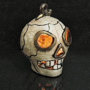 Puede incluir: Una escultura de cráneo de cerámica con un acabado de esmalte craquelado color crema y detalles metálicos oscuros. El cráneo presenta cuencas oculares huecas, una cavidad nasal triangular y una amplia sonrisa, iluminadas desde dentro por una luz naranja.