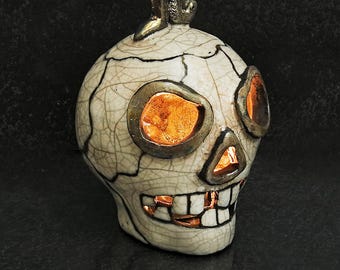 May include: A ceramic skull sculpture with a cream-colored crackle glaze finish and dark metallic accents. The skull features hollow eye sockets, a triangular nose cavity, and a wide grin, all illuminated from within by a warm orange light.