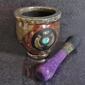 May include: A decorative mortar and pestle set. The mortar is brown with a silver rim and a circular design with a turquoise center. The pestle is purple with a black tip and silver accents. The set is designed for grinding herbs and spices.