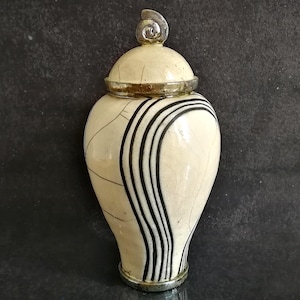 Może przedstawiać: Kremowy ceramiczny wazon z pokrywką i srebrną metalową obwódką. Wazon posiada czarne pionowe paski i spiralny wzór na pokrywce. Jest to przedmiot dekoracyjny.