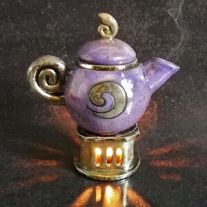 May include: A ceramic teapot in shades of purple and gold, with a decorative swirl design. The teapot sits on a small stand with lit candles, casting a warm glow. Steam rises from the spout, suggesting a warm beverage.