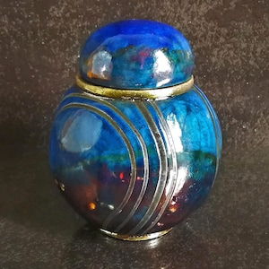 May include: A small, round, lidded jar featuring a glossy, multi-toned finish that transitions from deep blue at the top to a dark, reddish-brown at the base. The surface is decorated with a series of concentric, etched silver-colored lines.