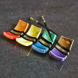 May include: A set of colourful chopstick rests in red, yellow, blue, and purple, each with black and silver stripes. Two chopsticks are placed on top. The items are on a dark, textured surface.