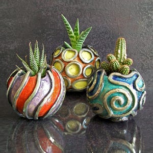 May include: Three decorative ceramic plant pots, each containing a succulent or cactus. The pots are spherical with distinct designs: one with orange and purple stripes, one with yellow circles, and one with blue swirls. The plants add a touch of green.