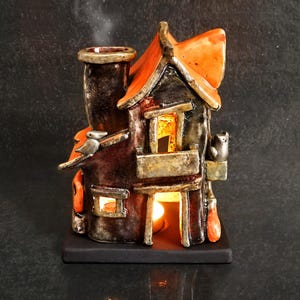 Pode incluir: Um queimador de incenso de cerâmica em forma de casa com acabamento em castanho escuro e preto, apresentando um telhado laranja brilhante, uma chaminé alta que emite fumo e uma pequena varanda. Uma luz quente brilha do interior da estrutura.