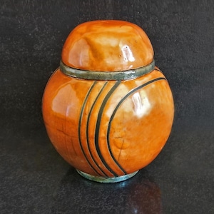 May include: A glazed orange ceramic jar with a lid. The jar has black curved lines and a green band around the middle. The jar is round and sits on a dark surface.