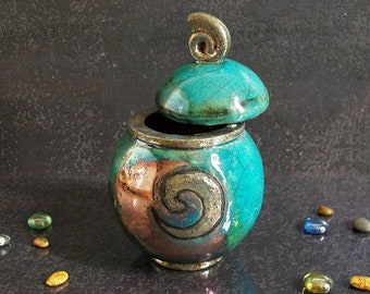 Puede incluir: Un tarro de cerámica de color verde azulado con tapa, decorado con un diseño de espiral dorado. El tarro está sobre una superficie gris oscuro.