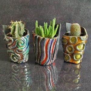 Peut inclure: Trois petits pots de fleurs en céramique avec des cactus et des plantes succulentes. Les pots sont décorés de motifs colorés et de formes géométriques. Les plantes sont vertes et épineuses, et les pots sont dans les tons de bleu, rouge, orange et jaune.