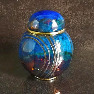 Könnte beinhalten: Ein rundes Keramikgefäß mit Deckel, das eine glänzende Oberfläche in tiefen Blau-, Violett- und Dunkelrottönen aufweist. Die Oberfläche ist mit drei geschwungenen, metallisch silbernen Linien und einem goldfarbenen Rand an der Öffnung verziert.