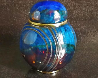 Könnte beinhalten: Ein rundes Keramikgefäß mit Deckel, das eine glänzende Oberfläche in tiefen Blau-, Violett- und Dunkelrottönen aufweist. Die Oberfläche ist mit drei geschwungenen, metallisch silbernen Linien und einem goldfarbenen Rand an der Öffnung verziert.