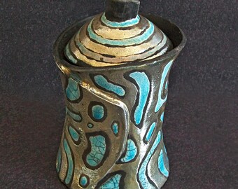 Może przedstawiać: Ręcznie robiony ceramiczny słoik z pokrywką. Słoik ma unikalny design z turkusowymi i srebrnymi akcentami. Pokrywka jest w paski w odcieniach beżu, niebieskiego i czarnego. Jest to element dekoracyjny, prawdopodobnie do przechowywania drobnych przedmiotów.