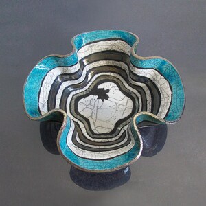 Può includere: Una ciotola decorativa in ceramica con una forma ondulata unica. La ciotola presenta un design sorprendente con bande alternate bianche, nere e grigie, accentuato da un bordo turchese. L'interno presenta un motivo floreale centrale.