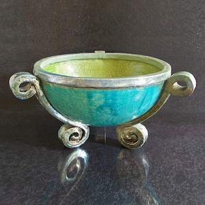 Può includere: Una ciotola in ceramica fatta a mano con esterno turchese e interno verde lime. La ciotola presenta manici decorativi in metallo e piedini arricciati. Il bordo è accentuato da una fascia argentata. Un pezzo decorativo unico.