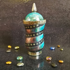May include: A decorative ceramic jar with a teal, copper and black swirl pattern and gold accents. The jar has a lid with a pointed top.