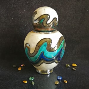 May include: A white ceramic jar with a blue, green, and gold wave design. The jar has a round lid with a similar wave design.