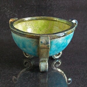May include: A handcrafted ceramic bowl with a turquoise exterior and a lime green interior. The bowl has three small feet and two handles. The rim is a dark brown color. The bowl is approximately 6 inches in diameter.